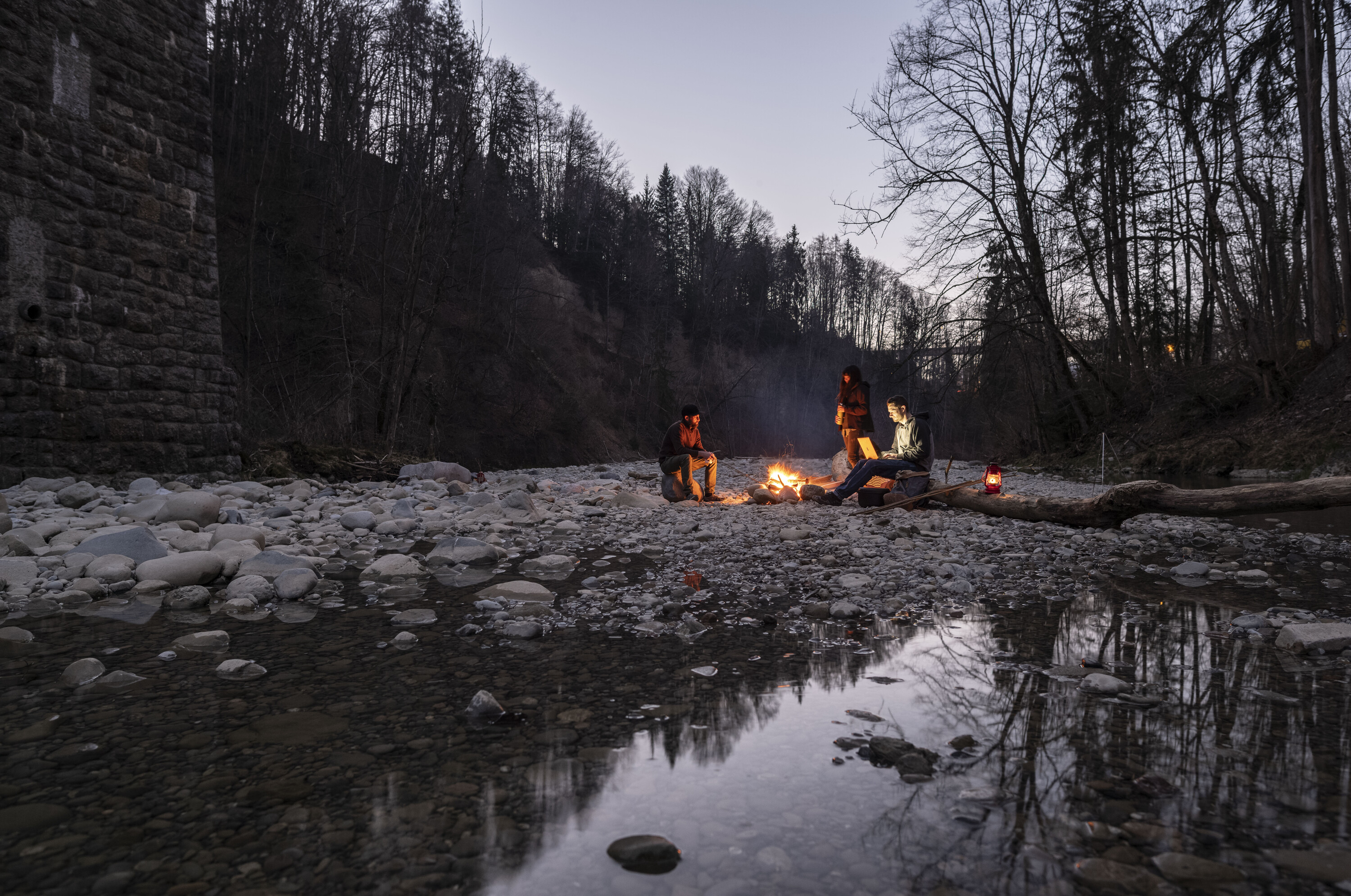 Headerbild: Personen sitzen um eine Feuerstelle in der Dämmerung. Eine Person hat ein Laptop dabei.